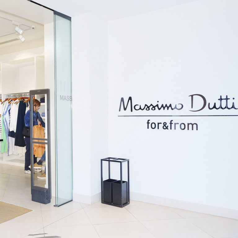 772741 - Outlet Social | Hablemos de Marketing Storefront of Massimo Dutti with a glass door and a shopper browsing clothes inside.