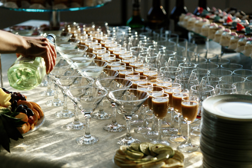 A hand pours champagne into a row of flute glasses on a catered reception table with fruit and desserts nearby.