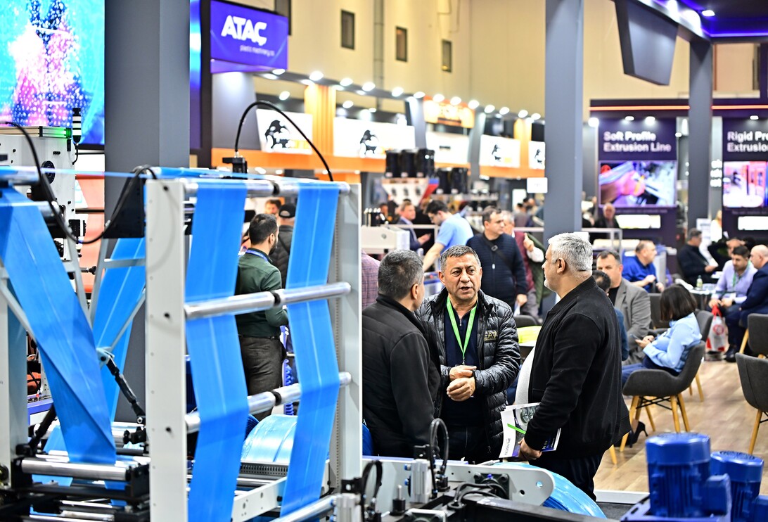 Group of men in dark jackets talking near a large blue textile machine at a trade show floor.
