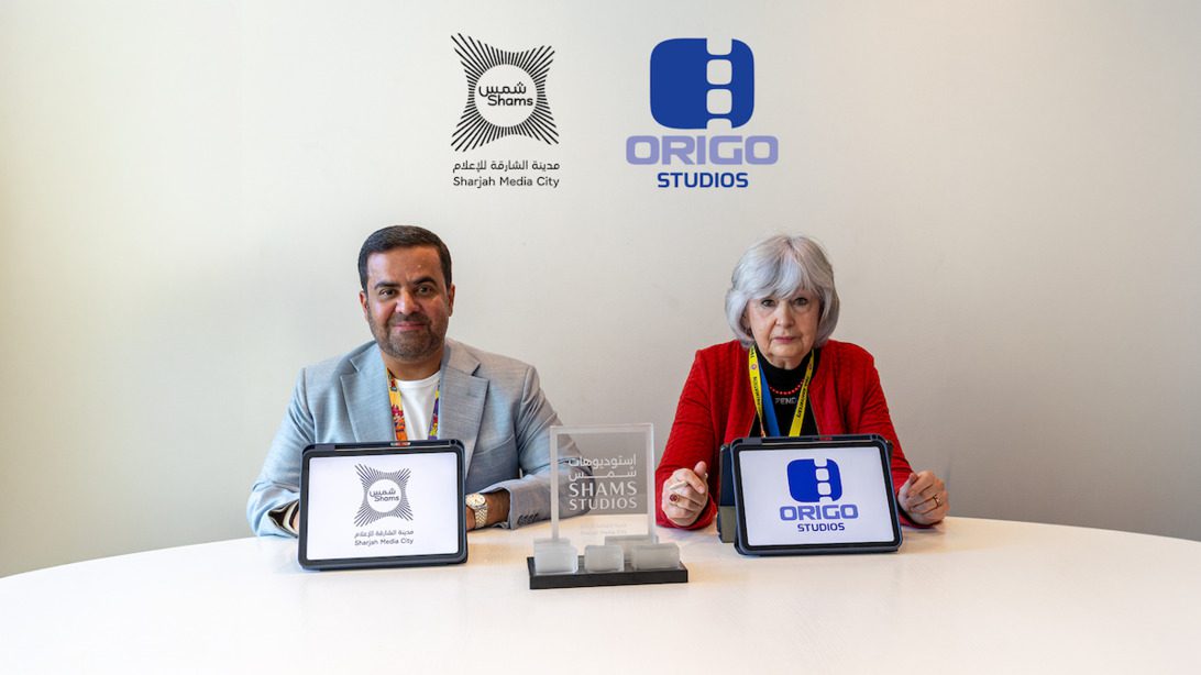 Two people sit at a white table with tablets displaying Shams and Origo Studios logos, indicating a collaboration, with the same logos on the wall behind them and a central trophy on the table.